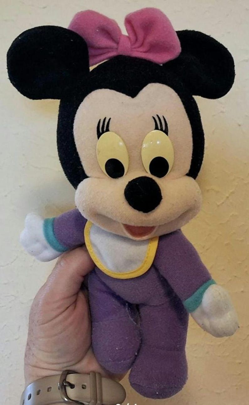 Pu&ograve; includere: Un peluche di un personaggio topo dei cartoni animati con orecchie nere, un fiocco rosa e grandi occhi gialli. Il personaggio indossa una tutina viola con polsini color ottanio e una bavaglino bianco con bordo giallo e mani bianche.