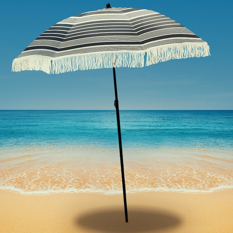 Puede incluir: Una sombrilla de playa a rayas blancas y negras con un borde de flecos blancos. La sombrilla est&aacute; de pie en una playa de arena con agua de mar azul en el fondo.