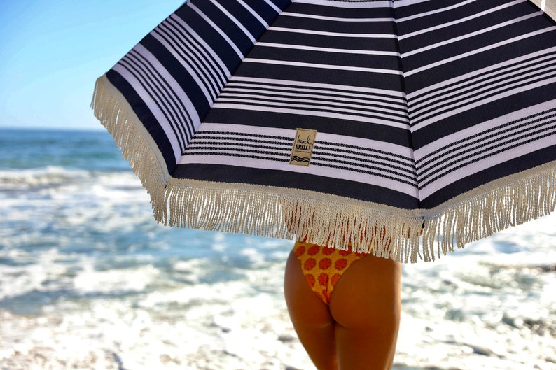 Puede incluir: Una sombrilla de playa a rayas blancas y negras con un borde de flecos blancos. La sombrilla tiene una peque&ntilde;a etiqueta marr&oacute;n con el texto "Beach Umbrella". La sombrilla est&aacute; delante de una persona que lleva la parte inferior de un bikini floral amarillo y naranja.