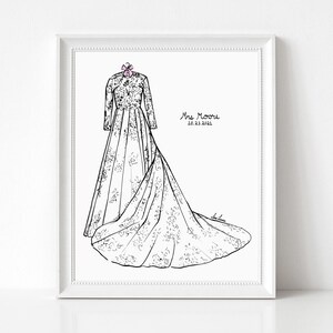 May include: A framed black and white line art illustration of a long-sleeved wedding dress with a floral pattern and a train, featuring a small pink bow at the neckline. The text Mrs Moore and 10.23.2021 are printed on the right side of the dress.