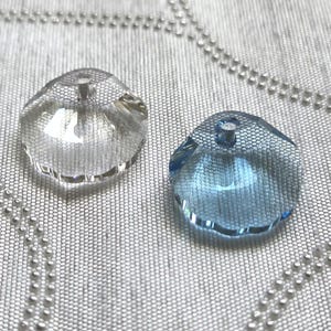 May include: Two faceted, clear and blue crystal pendants. The clear pendant is on the left, and the blue pendant is on the right. Both have a hole at the top for stringing. The pendants are on a textured, light gray surface.