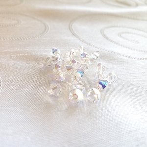 May include: A group of clear iridescent crystal beads, scattered on a white fabric background.