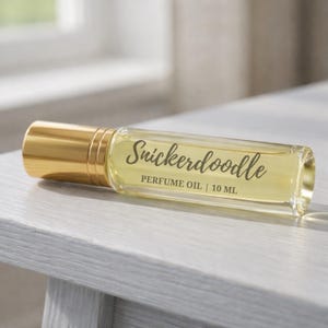 May include: A clear glass perfume oil bottle with a gold cap, labeled "Snickerdoodle PERFUME OIL | 10 ML". The bottle contains a light yellow liquid and rests on a light gray surface.