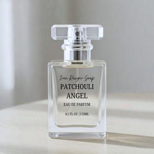 May include: A clear, rectangular perfume bottle with a silver atomizer and a faceted, rectangular cap. The bottle has the text "Iron Ranger Soap PATCHOULI ANGEL EAU DE PARFUM 4.5 FL OZ | 133ML" in black. The perfume bottle is on a light background.