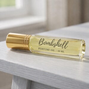 May include: A clear glass perfume oil bottle with a gold cap, labeled "Bombshell PERFUME OIL | 10 ML". The bottle contains a light yellow liquid and rests on a white wooden surface. The background is blurred.