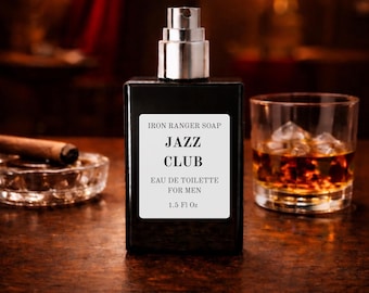 May include: A black rectangular bottle of "Jazz Club" eau de toilette with a silver atomizer. The label reads "Iron Ranger Soap" and "1.5 Fl Oz". A glass of amber liquid with ice cubes and a cigar in an ashtray are also visible.