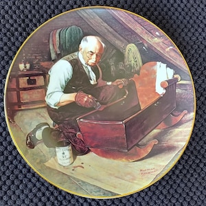 Puede incluir: Plato decorativo con una ilustración detallada de un hombre fabricando una cuna de madera. La escena está ambientada en un taller, con el hombre arrodillado y trabajando en la cuna. Borde dorado.