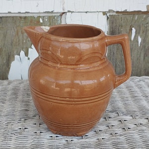 May include: A brown glazed ceramic pitcher with a wide mouth and a handle. The pitcher has a decorative design around the top and bottom.