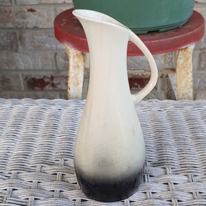 May include: A white ceramic vase with a black ombre design and a handle. The vase is sitting on a wicker surface.
