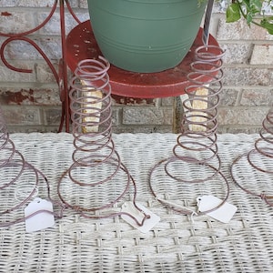 Op de afbeelding: Vijf roestige metalen veercoils, elk gevormd als een kegel, zijn gerangschikt op een wit rieten oppervlak. De coils worden gebruikt als plantenstandaards.