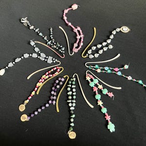 May include: A collection of beaded bookmarks in various colors and designs. Some feature gold-colored charms and decorative beads, while others have unique shapes like flowers and shells. The bookmarks are arranged in a circular pattern against a black background.