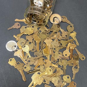 May include: A collection of vintage brass keys spilling from a clear glass jar onto a dark surface. The keys vary in size and design, with some showing signs of wear. The jar is partially filled with more keys, suggesting a large assortment.