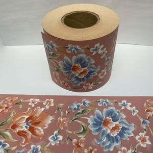 May include: A roll of floral wallpaper border featuring a repeating pattern of large blue and orange flowers with smaller white and blue blossoms on a dusty rose background. The wallpaper is unrolled to show the pattern.