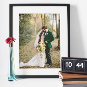 May include: A black framed photo of a bride and groom in a forest setting. The bride is wearing a white lace dress and the groom is wearing a green velvet suit. The photo is displayed on a white surface with a vase of red flowers and a black clock that reads 10:44.