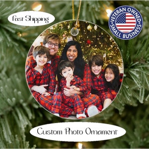 May include: A round, white ceramic Christmas ornament with a photo of a family of five wearing red and black plaid pajamas in front of a Christmas tree. The ornament has a gold string for hanging.