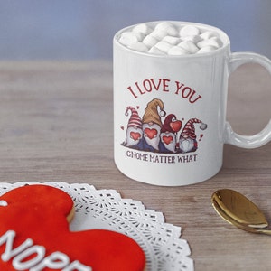 May include: White ceramic coffee mug with a red and white gnome design. The mug has the text "I love you gnome matter what" printed on it. The mug is filled with marshmallows.