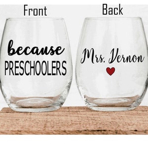 May include: Two clear stemless wine glasses with black text. The front glass reads "because PRESCHOOLERS". The back glass reads "Mrs. Vernon" with a red heart. The glasses are on a wooden surface.