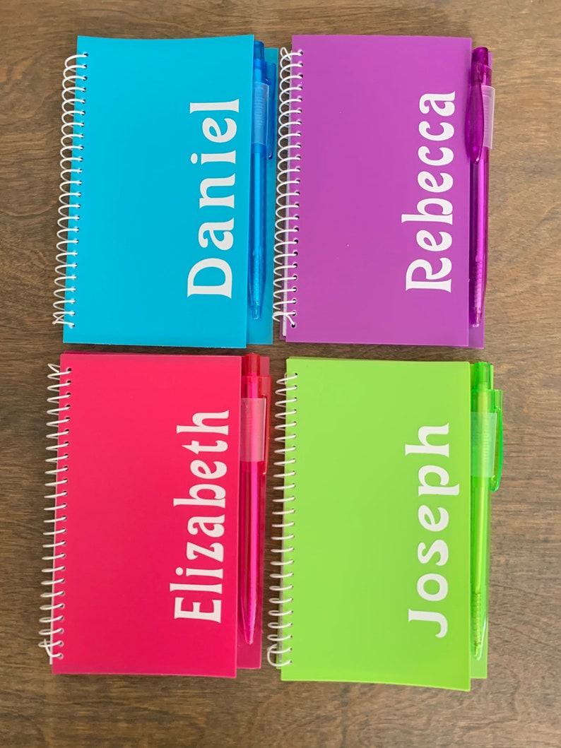 May include: Four colorful spiral notebooks with personalized names: Daniel, Rebecca, Elizabeth, and Joseph. Each notebook has a matching colored pen.
