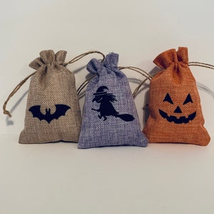 May include: Three small burlap bags with drawstring closures. Each bag features a black Halloween-themed design: a bat, a witch on a broom, and a jack-o'-lantern face. The bags are in shades of tan, purple, and orange.