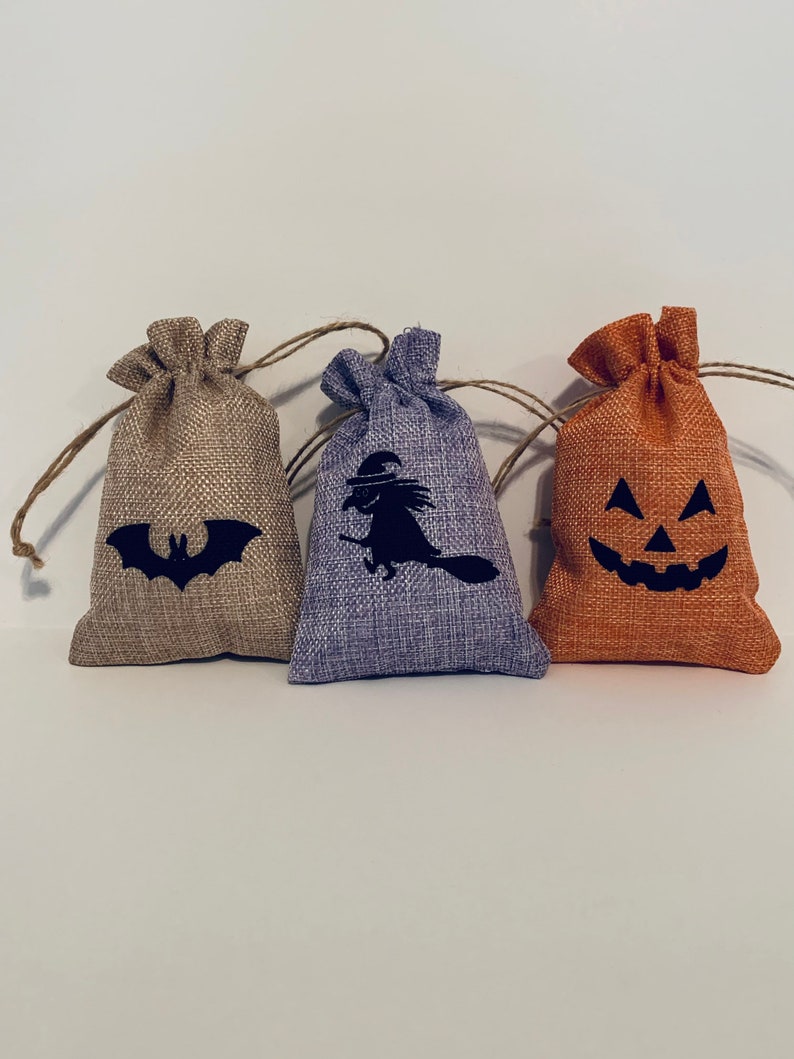 May include: Three small burlap bags with drawstring closures. Each bag features a black Halloween-themed design: a bat, a witch on a broom, and a jack-o'-lantern face. The bags are in shades of tan, purple, and orange.