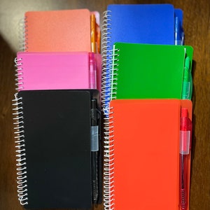 May include: A stack of six spiral bound notebooks with different colored covers. Each notebook has a pen attached to the spine. The colors of the notebooks are pink, black, orange, green, blue, and orange.