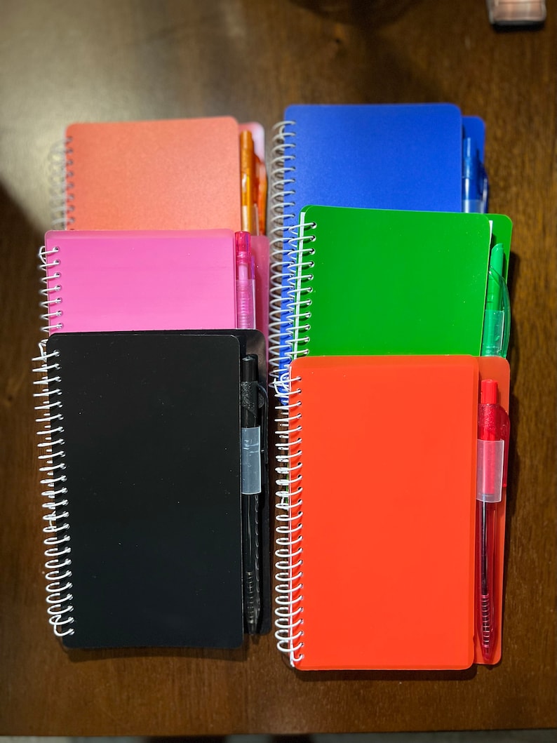 May include: A stack of six spiral bound notebooks with different colored covers. Each notebook has a pen attached to the spine. The colors of the notebooks are pink, black, orange, green, blue, and orange.