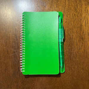 May include: A green spiral bound notebook with a green pen attached to the spine.