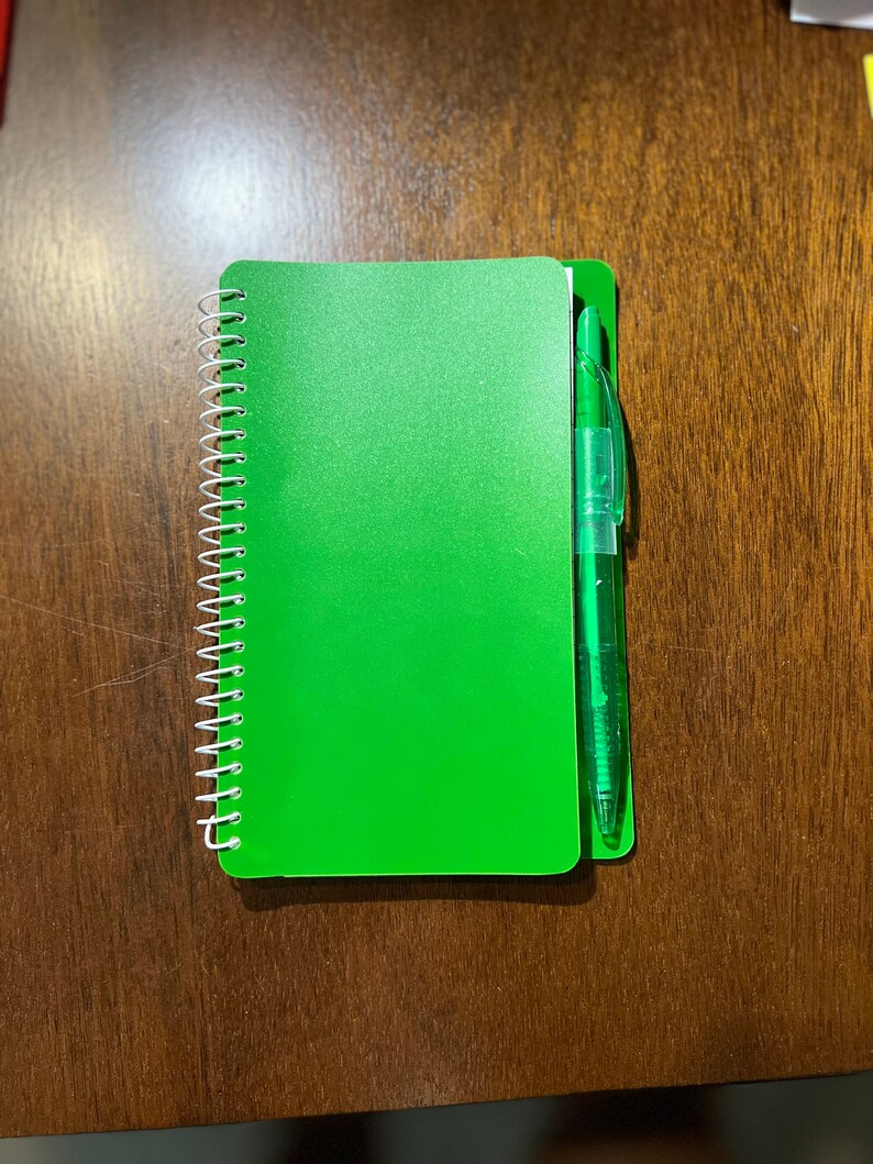 May include: A green spiral bound notebook with a green pen attached to the spine.