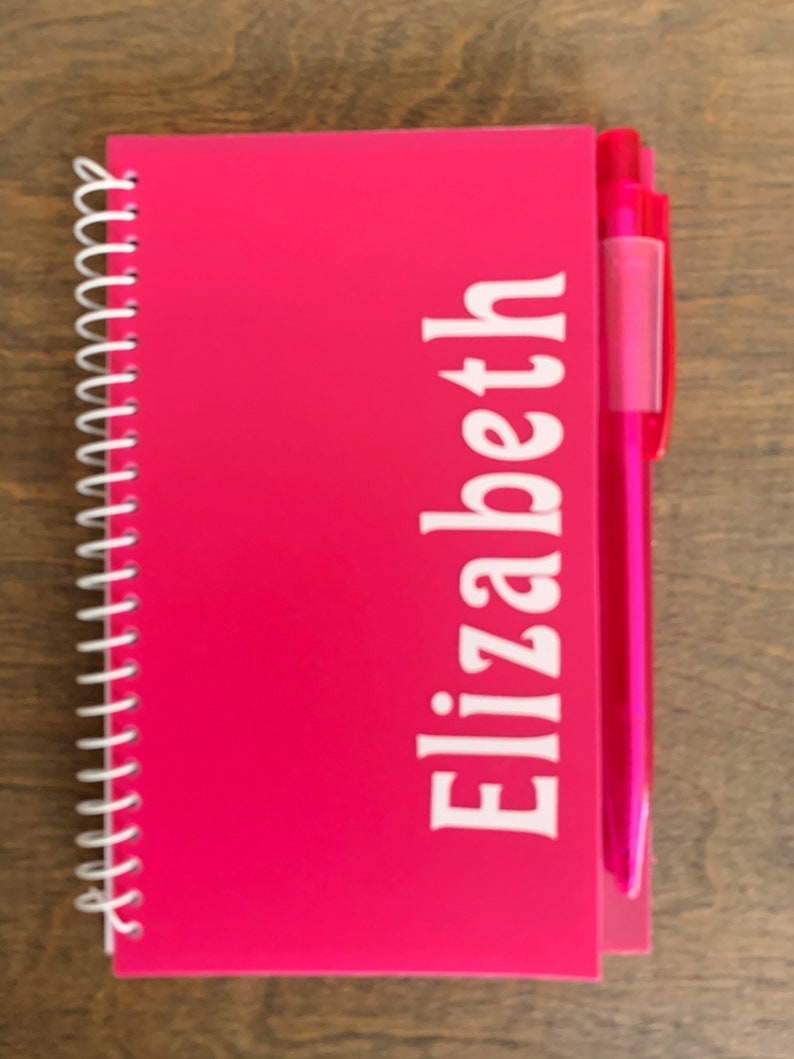 May include: A pink spiral bound notebook with the name "Elizabeth" printed in white on the front cover. A pink pen is attached to the notebook.
