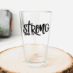 May include: A clear glass pint glass with a black vinyl decal that reads "strong" with a cross above the word and "Joshua 1:9" below. The glass is sitting on a wooden surface.