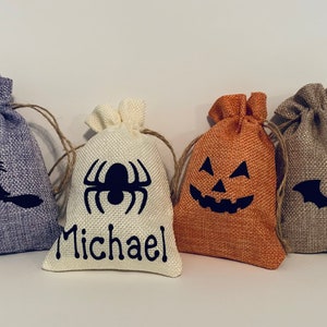 May include: Four small burlap drawstring bags with Halloween designs. The bags are in shades of purple, cream, orange, and tan. Each bag features a black graphic: a witch on a broom, a spider, a jack-o'-lantern face, and a bat. One bag has the name "Michael".