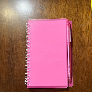 May include: A pink spiral bound notebook with a clear plastic cover and a pink pen attached to the side.