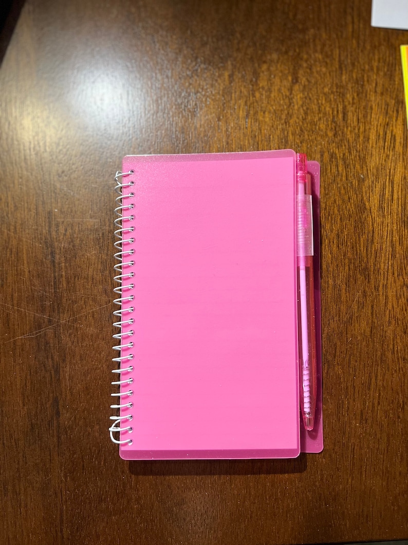 May include: A pink spiral bound notebook with a clear plastic cover and a pink pen attached to the side.