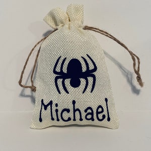 May include: A small, cream-coloured burlap drawstring bag featuring a dark blue spider design and the name "Michael". The bag has a textured surface and is tied with a brown string.