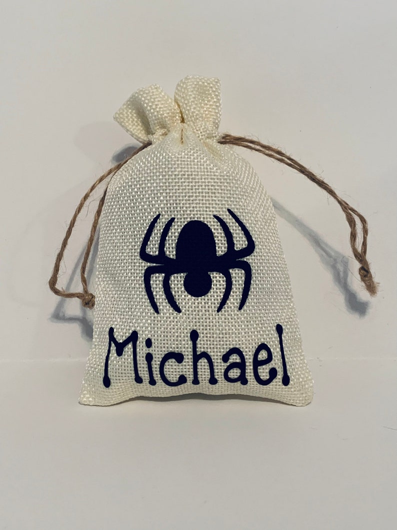 May include: A small, cream-coloured burlap drawstring bag featuring a dark blue spider design and the name "Michael". The bag has a textured surface and is tied with a brown string.