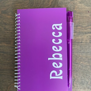 May include: A purple spiral bound notebook with the name "Rebecca" printed in white on the front cover. A purple pen is attached to the notebook.