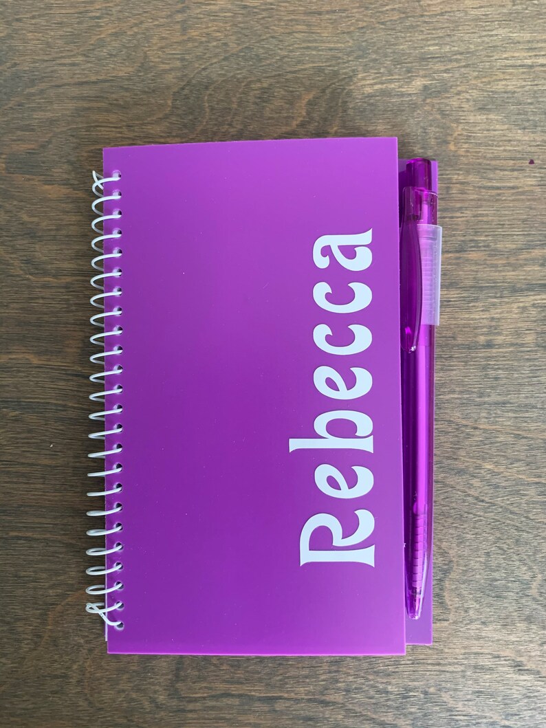May include: A purple spiral bound notebook with the name "Rebecca" printed in white on the front cover. A purple pen is attached to the notebook.