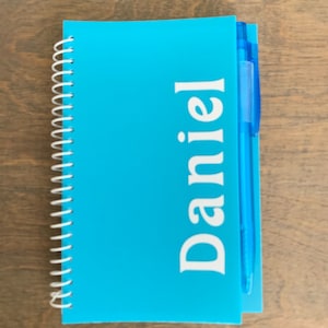May include: A blue spiral notebook with the name "Daniel" printed in white on the front cover. A blue pen is attached to the notebook.