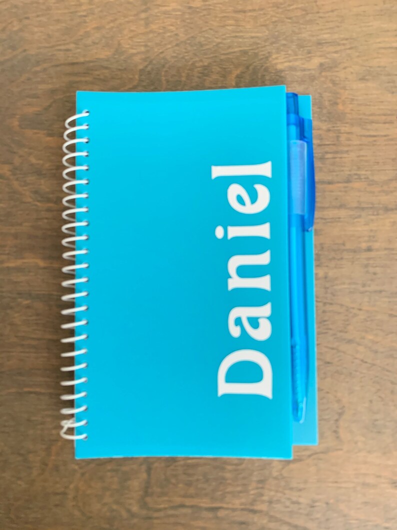 May include: A blue spiral notebook with the name "Daniel" printed in white on the front cover. A blue pen is attached to the notebook.