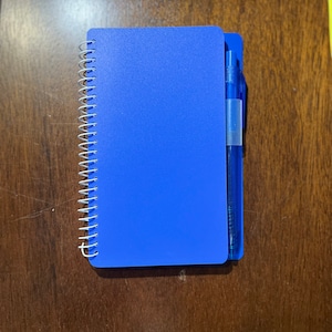 May include: A blue spiral bound notebook with a blue pen attached to the cover.