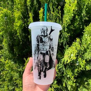 Può includere: Una tazza riutilizzabile bianca con un'immagine in bianco e nero di un personaggio di Mandalorian. La tazza ha una cannuccia verde e un coperchio trasparente.