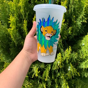 Op de afbeelding: Een witte herbruikbare beker met een doorzichtig plastic deksel en een rietje. De beker heeft een kleurrijk beeld van Simba uit The Lion King met een groene en blauwe manen.