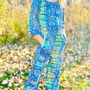 May include: A young person wearing a blue and green snake print jumpsuit with neon green trim. The jumpsuit has pockets on the sides. The person is standing in a grassy area with fallen leaves.