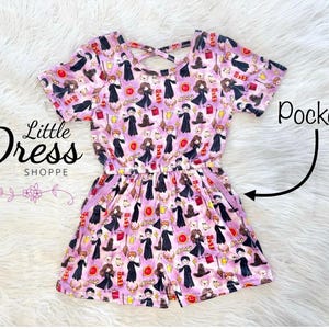 May include: A pink and purple short-sleeved dress with a Harry Potter theme. The dress has a criss-cross back and pockets on the sides. The dress is covered in a print of Harry Potter characters and objects.