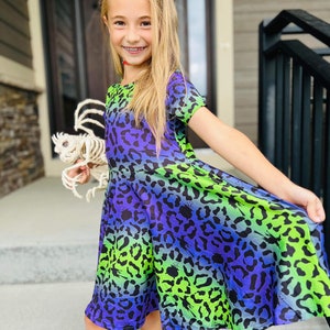 May include: A young girl wearing a green, blue, and purple leopard print dress. The dress has a short sleeve and a flared skirt. The girl is holding a skeleton prop in her hand.