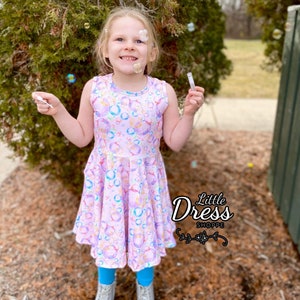 May include: A young girl wearing a pink sleeveless dress with a bubble print. The dress has a ruffled hem and is made of a soft, stretchy fabric. The girl is holding a bubble wand and blowing bubbles. The background is a blurred image of a green lawn and trees. The text "Little Dress Shoppe" is visible in the lower right corner of the image.