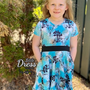 May include: A blue and white dress with a black waistband and a skull and cactus print. The dress has a ruffled hem and pockets on the sides. The dress is called "Little Dress Shoppe".