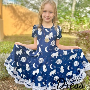 May include: A navy blue dress with a white wolf and celestial pattern. The dress features short sleeves, a round neckline, and a flared skirt with white lace trim. The text "Little Dress Shoppe" is visible on the bottom right.
