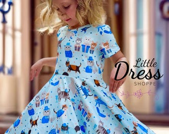 Peut inclure: Une robe bleu clair avec un motif de chats portant des chapeaux et tenant des cadeaux. La robe a une jupe ample et des manches courtes. Le texte "Little Dress Shoppe" est visible dans le coin inférieur droit de l'image.