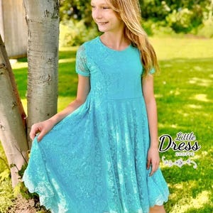 Puede incluir: Una niña pequeña con un vestido de encaje azul claro con un dobladillo festoneado. El vestido tiene un estampado floral y mangas cortas. La niña está de pie frente a un árbol y hierba verde.